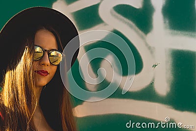 The hipster young girl wearing fashionable sunglasses and black hat with a graffiti as background Stock Photo
