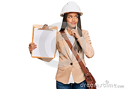 Beautiful hispanic woman architect showing black space on clipboard serious face thinking about question with hand on chin, Stock Photo