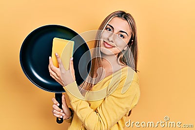 Beautiful hispanic woman cleaning cooking pan with scourer relaxed with serious expression on face Stock Photo