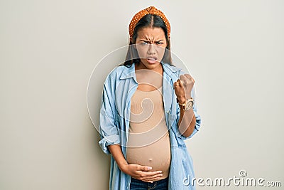 Beautiful hispanic woman expecting a baby, touching pregnant belly angry and mad raising fist frustrated and furious while Stock Photo