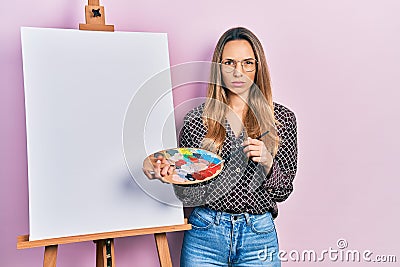 Beautiful hispanic woman standing drawing with palette by painter easel stand skeptic and nervous, frowning upset because of Stock Photo