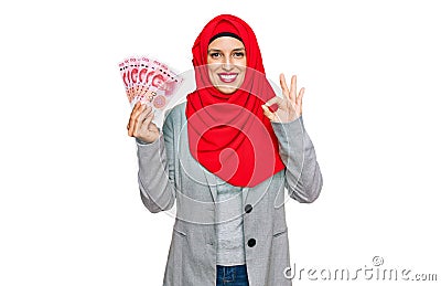 Beautiful hispanic woman wearing islamic hijab holding chinese yuan banknotes doing ok sign with fingers, smiling friendly Stock Photo