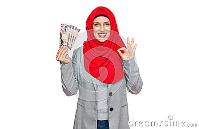 Beautiful hispanic woman wearing islamic hijab holding japanese yen banknotes doing ok sign with fingers, smiling friendly Stock Photo