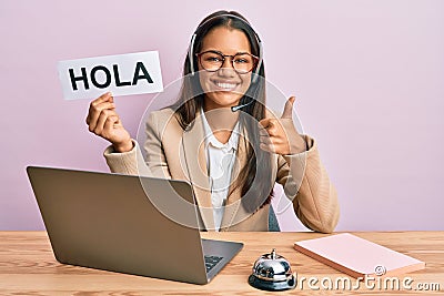 Beautiful hispanic woman wearing operator headset holding hola greenting smiling happy and positive, thumb up doing excellent and Stock Photo