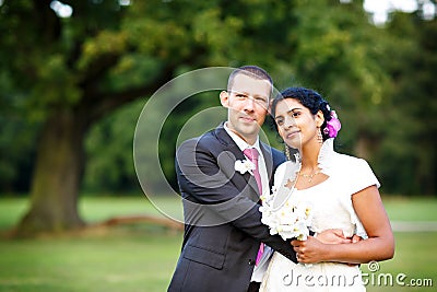 Beautiful indian bride and caucasian groom, in summer park. Happy young woman holding flowers. Young man smiling, Couple Stock Photo