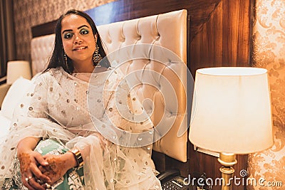 A beautiful Indian bride in traditional wear waiting in Hotel room next to lamp. A smiling girl wearing jewellery and designer Stock Photo