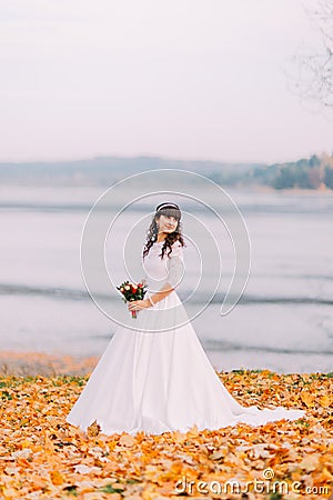 Beautiful innocent thoughtful bride in gorgeous white dress stands on fallen leaves at riverside Stock Photo