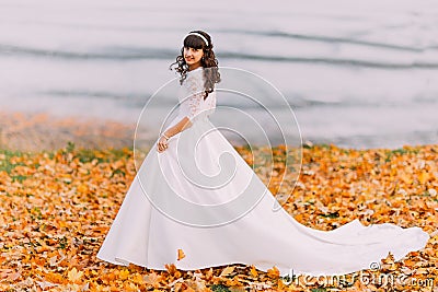 Beautiful innocent young brunette bride in gorgeous white dress stands on fallen leaves at riverside Stock Photo