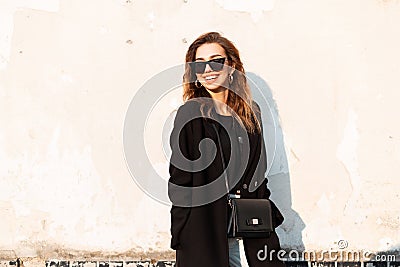 Beautiful joyful young hipster woman with a smile in a black luxurious coat in trendy dark sunglasses with a leather bag in jeans Stock Photo