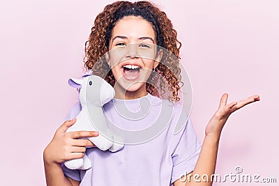 Beautiful kid girl with curly hair holding animal doll toy celebrating victory with happy smile and winner expression with raised Stock Photo