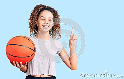 Beautiful kid girl with curly hair holding basketball ball surprised with an idea or question pointing finger with happy face, Stock Photo