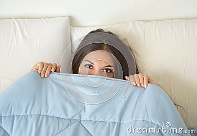 Beautiful lazy young woman lying down in the bed and sleeping. Teen girl with open eyes covers her face with blanket in the mornin Stock Photo