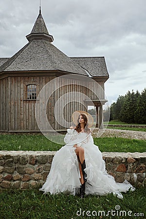 Beautiful leggy model girl in a wedding dress and straw hat posing near the wooden church in the countryside. Young Stock Photo