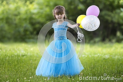 Beautiful little blond long-haired girl in nice long blue evening dress looking like princess looks in camera holding colorful bal Stock Photo