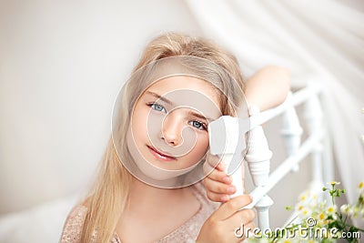 Beautiful little blonde girl with long hair sits on a bed with a bouquet of daisies in a bright white bedroom with a rustic interi Stock Photo