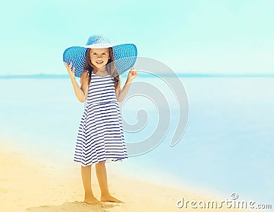 Beautiful little girl in a striped dress and summer straw hat relaxing on the beach near sea Stock Photo