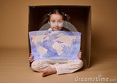 Beautiful little girl with two ponytails sitting inside a cardboard box with a poster- a painted image with World map, Stock Photo