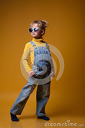 Beautiful little girl wearing stylish denim overall, yellow roll neck jumper and colorful sunglasses Stock Photo