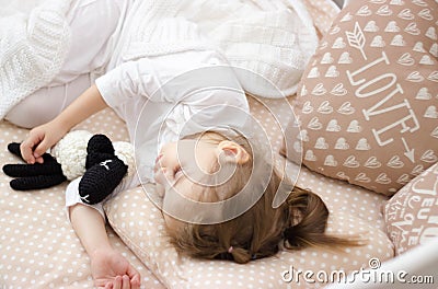 Beautiful little girl in white pajamas sleeps sweetly in a children& x27;s bed. She is hugging a handmade wool knitted sheep. Stock Photo