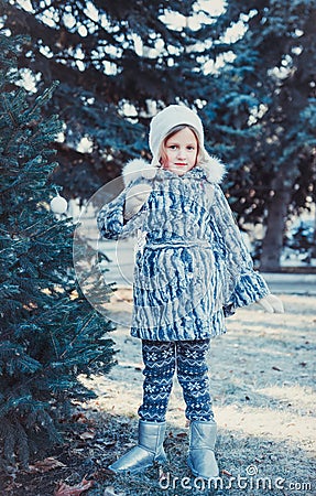 The beautiful little girl in winter wood. The girl is dressed in a gray fur coat. She is holding a white Christmas ball Stock Photo