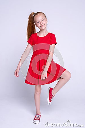 Beautiful little redhead girl in red dress and sneakers posing like model on white background. Stock Photo