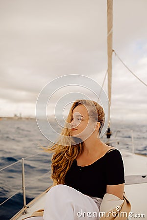 A beautiful long-haired girl sits on the bow of a barred yacht and enjoys life. The blonde is resting in the open sea or ocean. Stock Photo