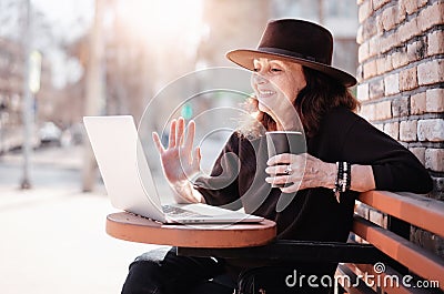 Beautiful mature elderly woman in hat making video call using laptop in street cafe on sunny day Stock Photo