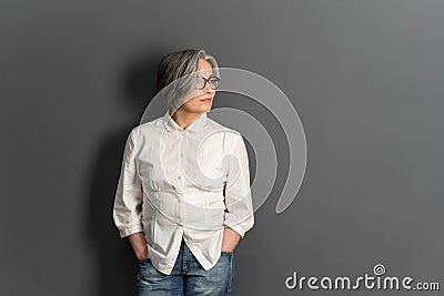 Beautiful mature lady in glasses stands hands in pockets looking at side. Elegant Gray-haired woman in whiite shirt on Stock Photo