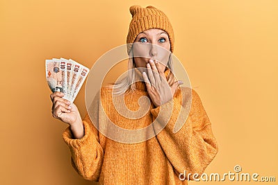 Beautiful middle age blonde woman holding 10 united kingdom pounds banknotes covering mouth with hand, shocked and afraid for Editorial Stock Photo