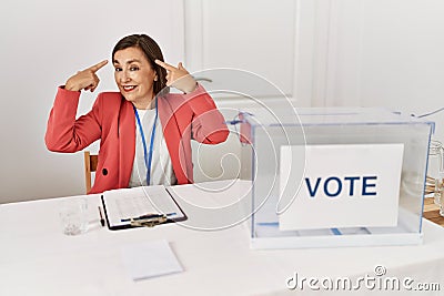 Beautiful middle age hispanic woman at political election sitting by ballot smiling pointing to head with both hands finger, great Stock Photo