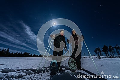 Beautiful middle aged men and women in snowshoes stand in night rare snowy winter forest under full moon light. Night walk Lapland Stock Photo