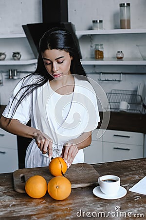 Beautiful mixed race girl in white robe preparing orange juice in morning and cutting oranges Stock Photo