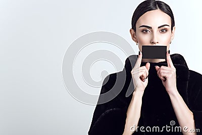 Beautiful model with sleek low ponytail wearing dark mink fur coat and holding black blank visit card in front of her mouth Stock Photo