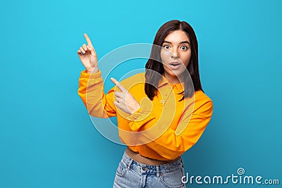 Beautiful multiethnicity woman in orange trendy shirt happily surprised expression pointing up against blue background Stock Photo