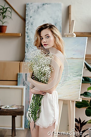 beautiful naked young woman holding white flowers and looking at camera in art studio Stock Photo