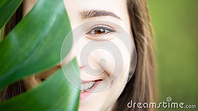 Beautiful natural young woman closeup behind big monstera leaf with green background in the woods Stock Photo