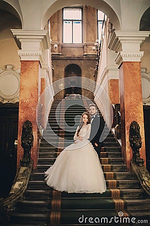 Beautiful newlyweds hugging on stairs the ancient house. Wedding portrait of a stylish groom and a young bride inside Stock Photo