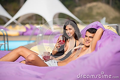Beautiful pair - guy and girl relaxing near swimming pool on cushioned loungers with drinks at the luxury resort Stock Photo