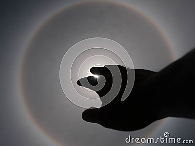 Beautiful photograph of the sun with a circular rainbow surrounded by a bright sky and white clouds with shadows of hands reaching Stock Photo