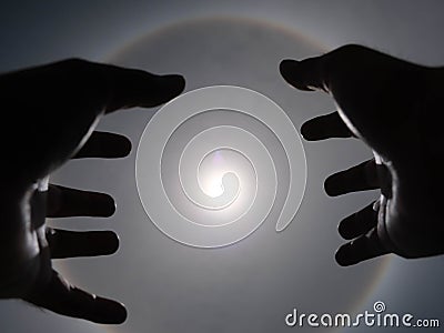 Beautiful photograph of the sun with a circular rainbow surrounded by a bright sky and white clouds with shadows of hands reaching Stock Photo