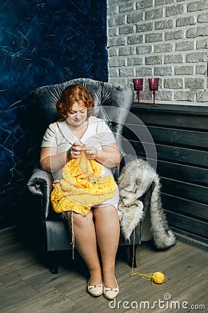 A beautiful plump redhead young woman with freckles sits in an armchair and knits a yellow sweater Stock Photo