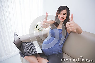 Beautiful pregnant asian woman sitting on a sofa with laptop on Stock Photo