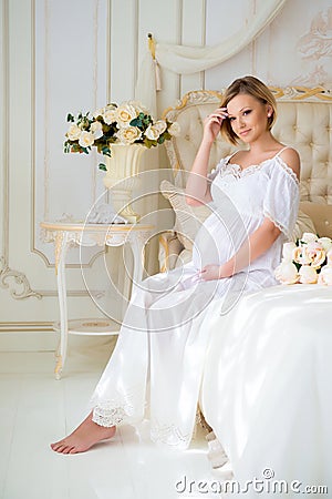 Beautiful pregnant girl in a home clothes sitting in the interior on a bed of roses Stock Photo