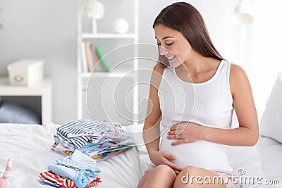 Beautiful pregnant woman with baby clothes on bed at home Stock Photo