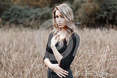 Beautiful pretty sexy young woman in a black stylish T-shirt with blond hair in trendy jeans poses in a field on a warm autumn day Stock Photo