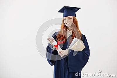 Beautiful redhead female graduate smiling holding books and diploma over white background. Stock Photo