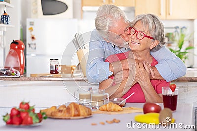 Beautiful retired senior couple having breakfast at home, starting morning with a kiss. Hot cappuccino croissant on the table Stock Photo