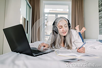 Beautiful senior concentrated businesslady in headphones, writing notes while watching video online webinar, learning or Stock Photo