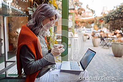 Beautiful senior woman uses videochat on laptop sitting on bench near cafe Stock Photo