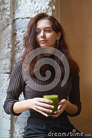 Beautiful serious stylish fashionable smart girl is sitting in cafe and drinking healthy green smoothie or latte vegan. Charming Stock Photo